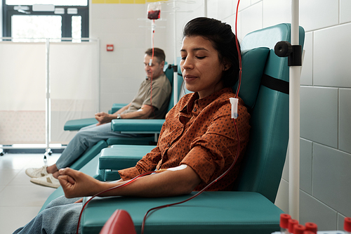 Young female and mature male donors giving blood for sick patients of modern hospital while sitting in armchairs in large ward