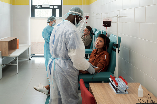 Young Hispanic female donor looking at African American clinician in protective coveralls, gloves, face screen and headband