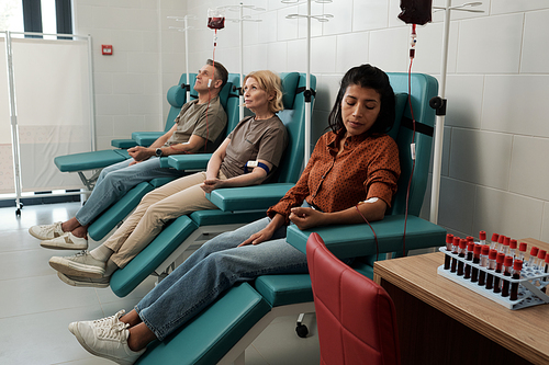Group of blood donors with dropper tubes sitting in row on chairs in large hospital ward during procedure of hemotransfusion
