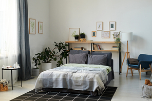 Part of spacious bedroom with double bed and shelves in the center surrounded by pictures in frames on walls and domestic plants