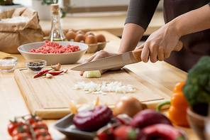 Hands of young female chopping fresh onion on wooden board while preparing italian pasta with vegetables and minced meat