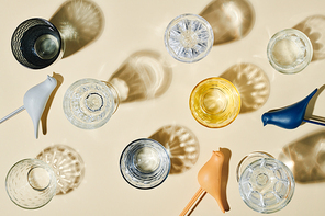 Several glasses containing clean water, their shadows and three plastic birds of blue, white and beige color over pastel background