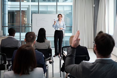 Young smiling female lecturer questioning audience after presentation of financial fluctuation graphs at seminar or conference