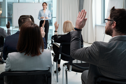 Several contemporary brokers raising hands to ask questions of business coach while sitting on chairs in rows in conference hall