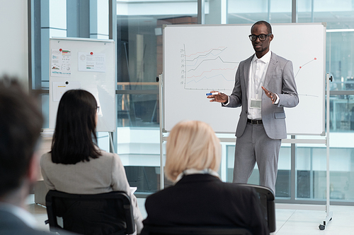 Confident teacher in elegant grey suit explaining audience graphic data on whiteboard during lecture of economics in conference hall