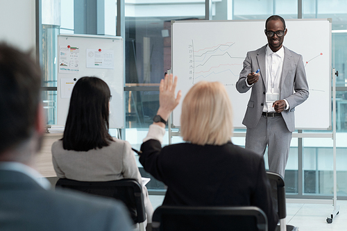 Young successful business coach in suit pointing at woman with raised hand at seminar while questioning audience after presentation