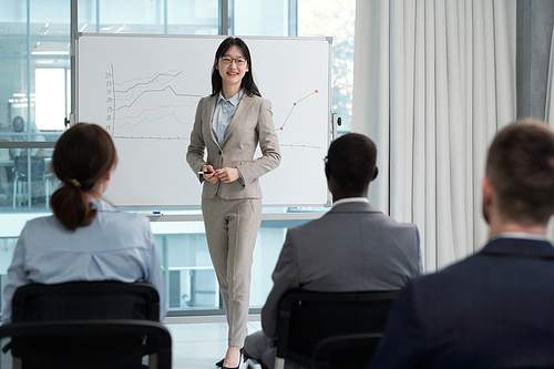 Happy young Asian female speaker presenting financial information to colleagues at business seminar or training in lecture hall