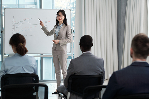 Successful business coach pointing at graphs on whiteboard at seminar while making presentation of fluctuations to colleagues