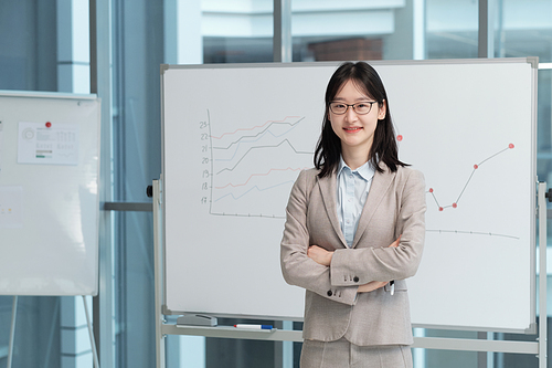 Young smiling female lecturer in formalwear and eyeglasses looking at camera while standing by whiteboard with financial graphs