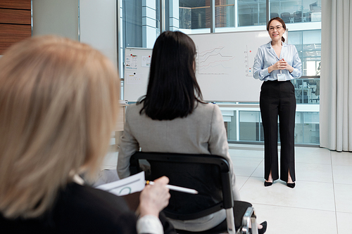 Young smiling business coach in formalwear communicating with audience at seminar while standing by whiteboard with graphs