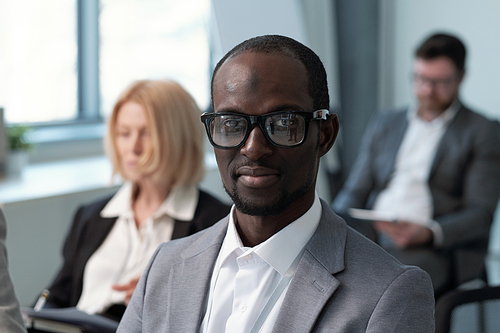 Young contemporary black man in formalwear and eyeglasses looking at camera while sitting against male and female colleagues