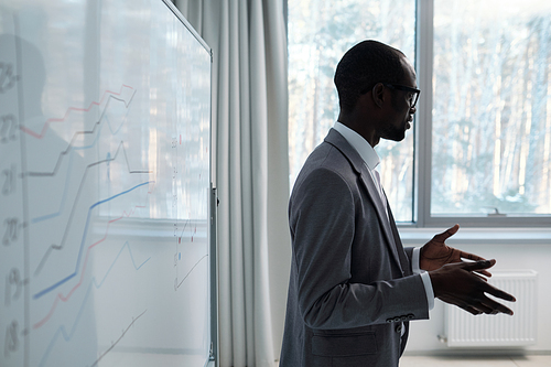 Young confident black man in grey suit explaining information to audience while standing by whiteboard with graphs