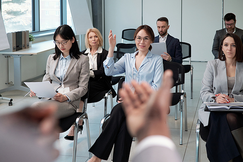 Young female speaker raising hand to ask question to business coach about subject of presentation while sitting among colleagues