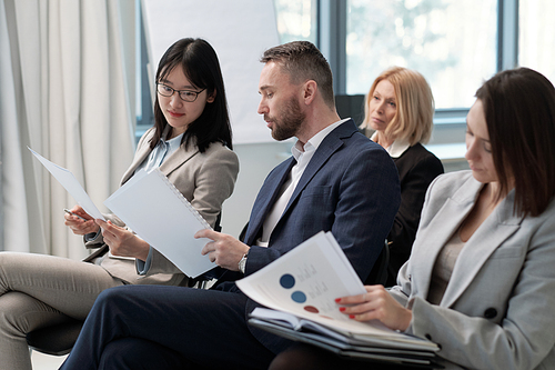 Young businessman in formalwear showing his report to Asian female colleague at conference while getting ready for presentation