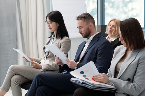 Group of serious brokers in formalwear looking through their reports at conference in lecture hall while getting ready for speech