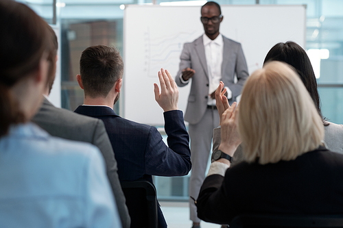 Rear view of businessman raising hand to answer question of coach while sitting among colleagues in front of African American speaker