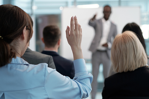 Hand of young female employee wishing to answer question of lecturer while sitting behind group of other speakers at conference