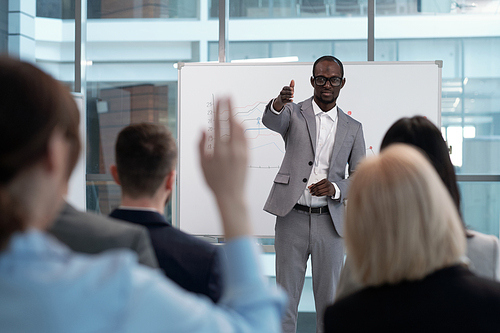 Young African American male business coach pointing at one of colleagues with raised hand wishing to answer his question