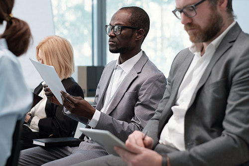 Young serious African American businessman with document listening to speaker while sitting between male and female colleagues