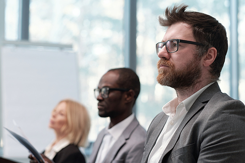 Young serious bearded businessman looking at speaker during presentation against two intercultural colleagues