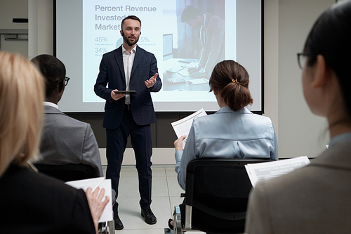 Young confident broker in elegant suit making presentation by interactive board while standing in front of audience