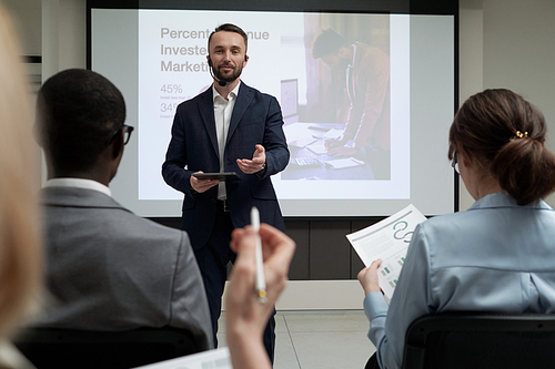 Contemporary young male economist in formalwear speaking in microphone by his mouth during presentation for colleagues