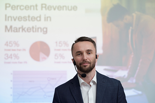 Confident young businessman with microphone by his face looking at camera against interactive board with financial information