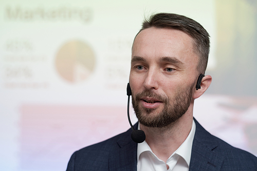 Young confident broker or business coach with microphone by his mouth making speech in front of audience at conference or seminar