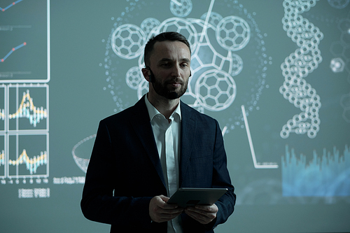 Serious male broker in formalwear holding tablet while making report to audience by interactive board at conference or seminar