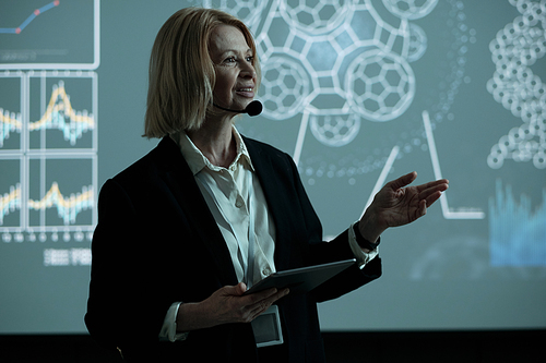 Happy blond mature businesswoman with tablet making speech at conference while standing against interactive board