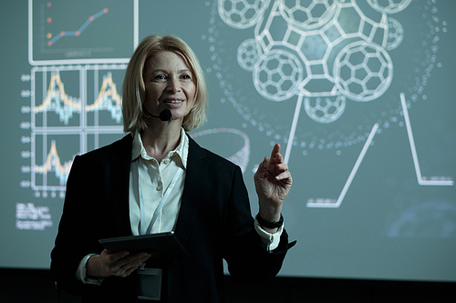 Confident mature female speaker with tablet and microphone by mouth standing by interactive board and making report for audience