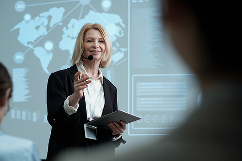 Happy mature female lecturer with tablet speaking to one of colleagues in audience while making report or presentation at conference