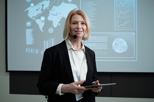 Happy mature female broker with tablet looking at camera against interactive board with data while making report for audience