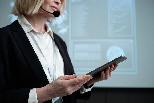 Contemporary female business coach with digital tablet making speech at conference while standing against interactive board with data