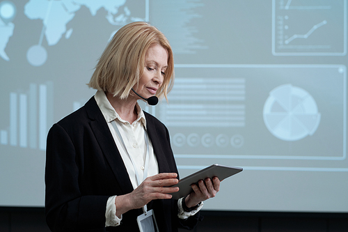 Serious female speaker or professor of economics with tablet reading lecture in auditorium while standing against interactive board