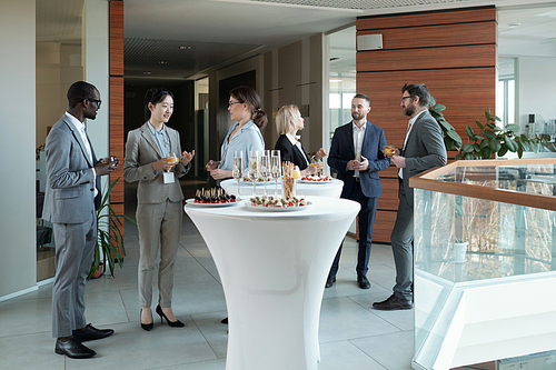 Large group of intercultural people in formalwear communicating at buffet while standing by tables served with snacks and champagne