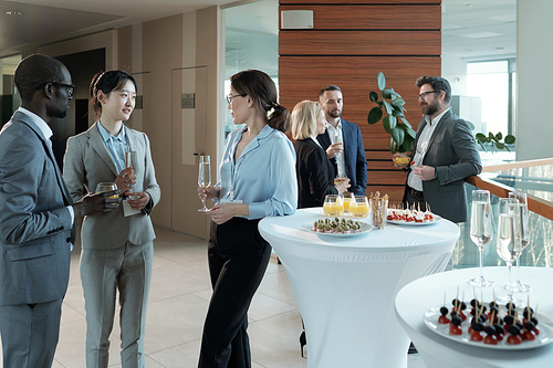 Young intercultural entrepreneurs with champagne discussing best reports after conference while standing by served tables