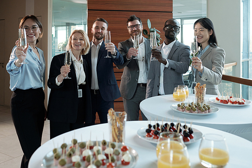 Group of cheerful business team in formalwear toasting at buffet in restaurant while standing by tables served with snacks and drinks