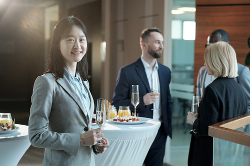 Happy young Asian businesswoman in formalwear holding flute of champagne and looking at camera against colleagues