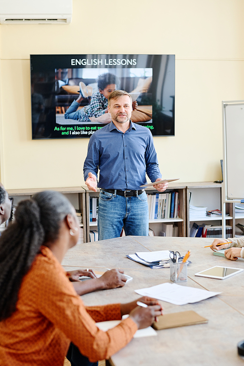 Vertical shot of mature Caucasian man standing at desk in front of multi-ethnic students teaching them English language