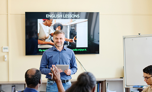 Mature Caucasian teacher holding digital tablet standing against screen with presentation on it working with migrant students
