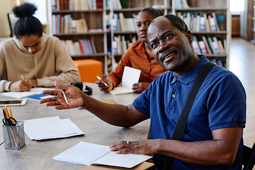 Portrait of mature Black man sitting at table in library asking teacher question about difficult task during lesson for immigrants
