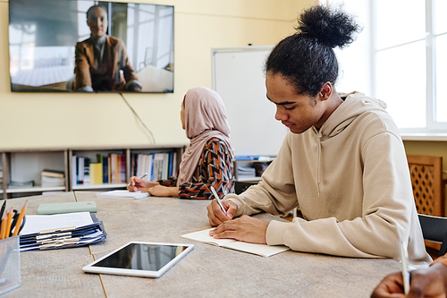 Multi-ethnic students sitting at table watching educational video and making notes in notebooks during lesson