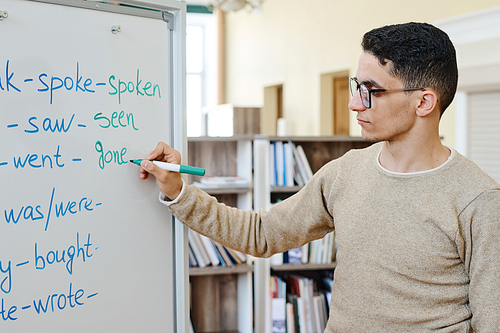 Handsome young Middle Eastern migrant student standing at whiteboard doing task with irregular verbs