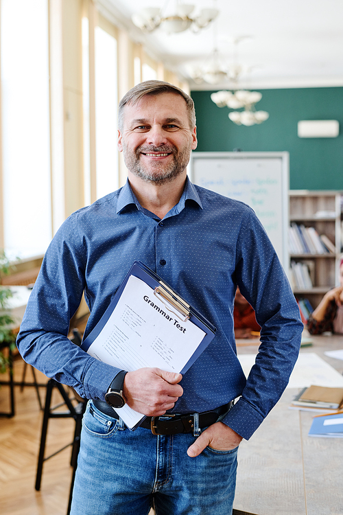 Vertical medium shot of joyful mature Caucasian man teaching English language holding clipboard with grammar tests smiling at camera