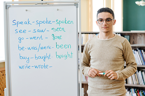 Horizontal medium portrait of young Middle-Eastern student standing at whiteboard with irregular verbs written on it looking at camera