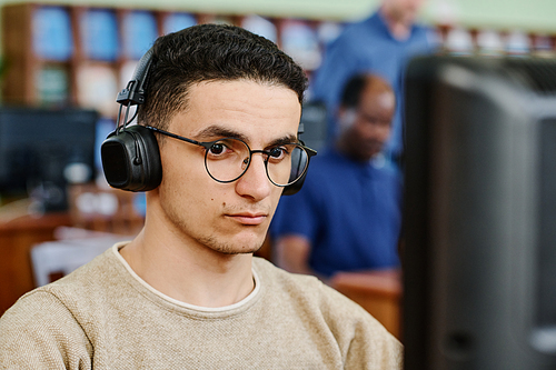 Young serious Middle Eastern studio wearing eyeglasses and headphones doing listening task during English lesson