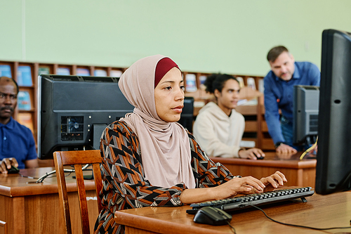 Portrait of modern Muslim woman wearing hijab sitting in university library working on desktop computer