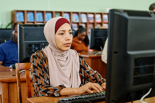 Portrait of pensive Muslim woman wearing hijab sitting in university library texting something on desktop computer