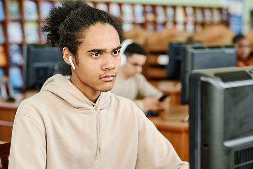 Young gen Z Black man wearing earphones sitting at desk in university library working on desktop computer and listening to music